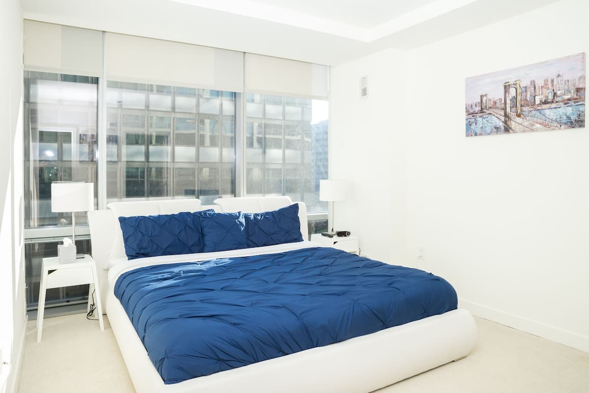 A modern hotel room with a large bed, white nightstands, and a cityscape painting on the wall.