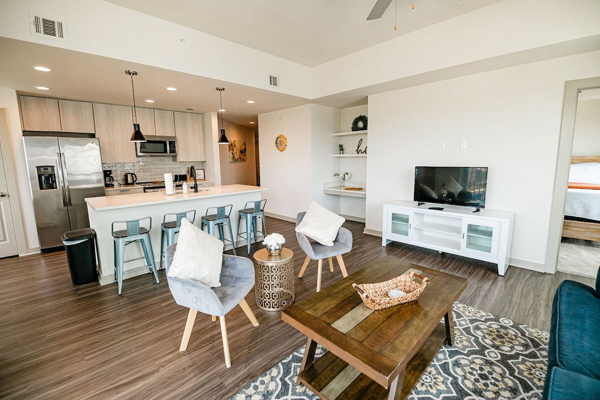 A modern living room with a wooden coffee table, two grey armchairs, a TV, and a kitchen area with a fridge, microwave, and bar stools.