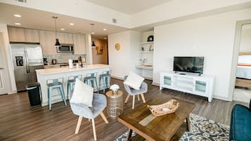 Un salon moderne avec une table basse en bois, deux fauteuils gris, un téléviseur et un coin cuisine équipé d’un réfrigérateur, d’un micro-ondes et de tabourets de bar.