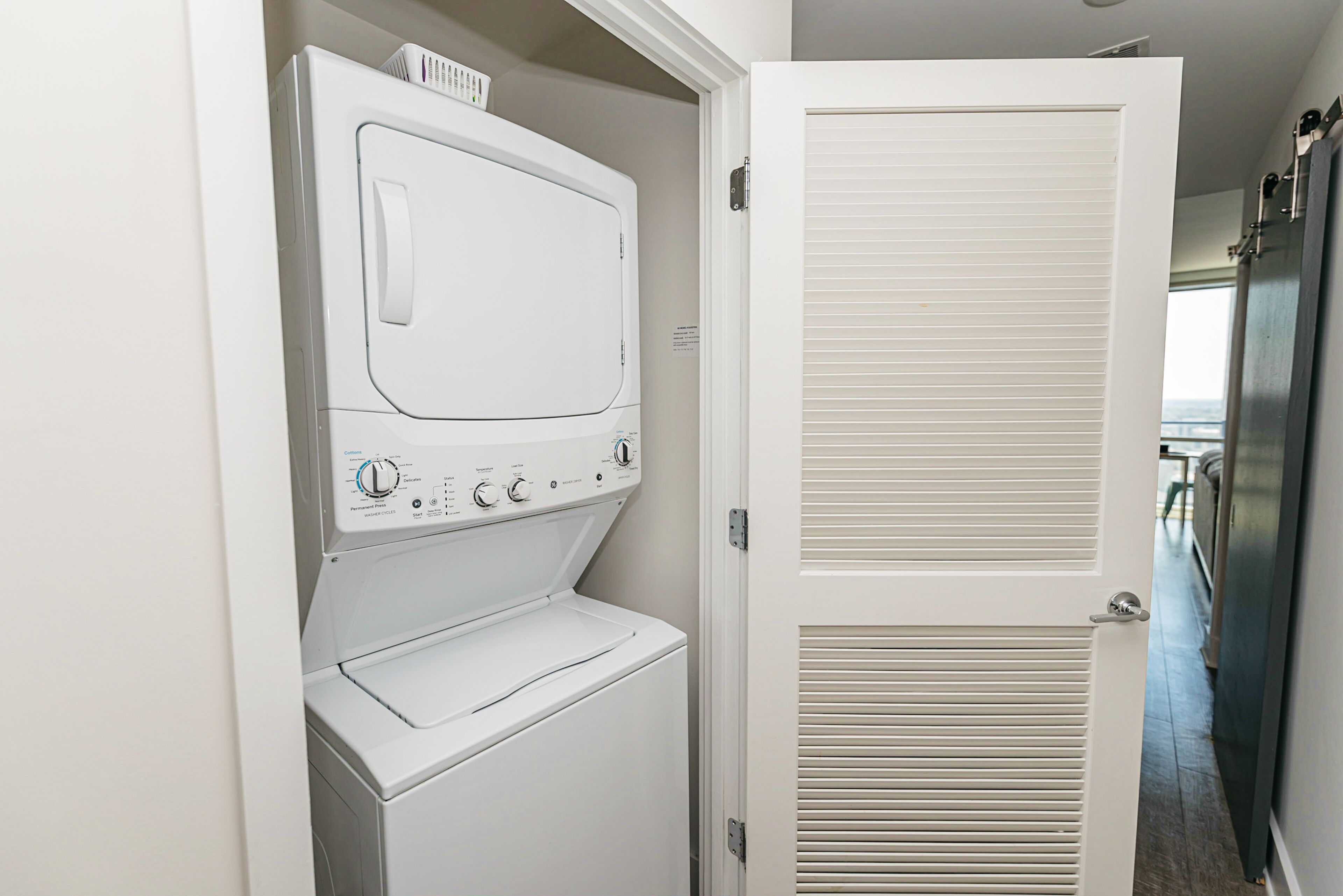 Een wasserette met een voorlader wasmachine en droger, een deur met verstelbare ventilatieopeningen, en een balkon in de achtergrond.