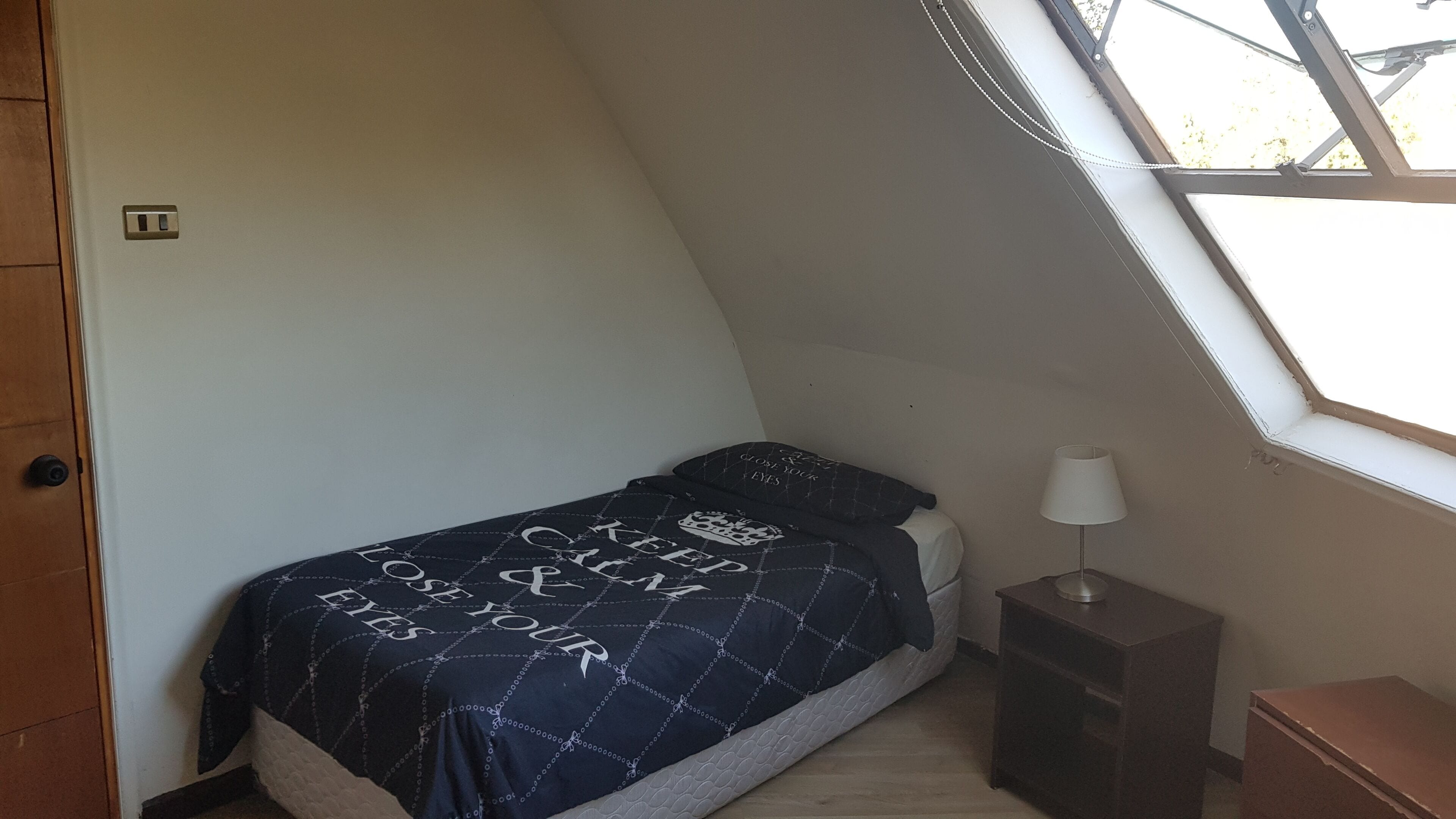 A small, sloped bedroom with a bed, a nightstand, a lamp, and a window.