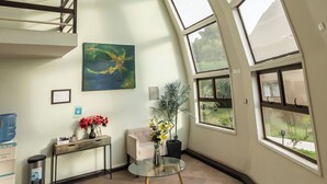 Una habitación amplia con techo alto, ventanas de tragaluces, una escalera, un cuadro, una pequeña mesa con flores y un cesto de basura.