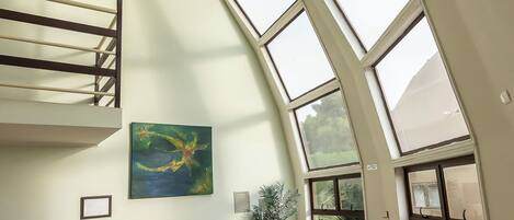 A spacious room with a high ceiling, skylight windows, a staircase, a painting, a small table with flowers, and a trash bin.