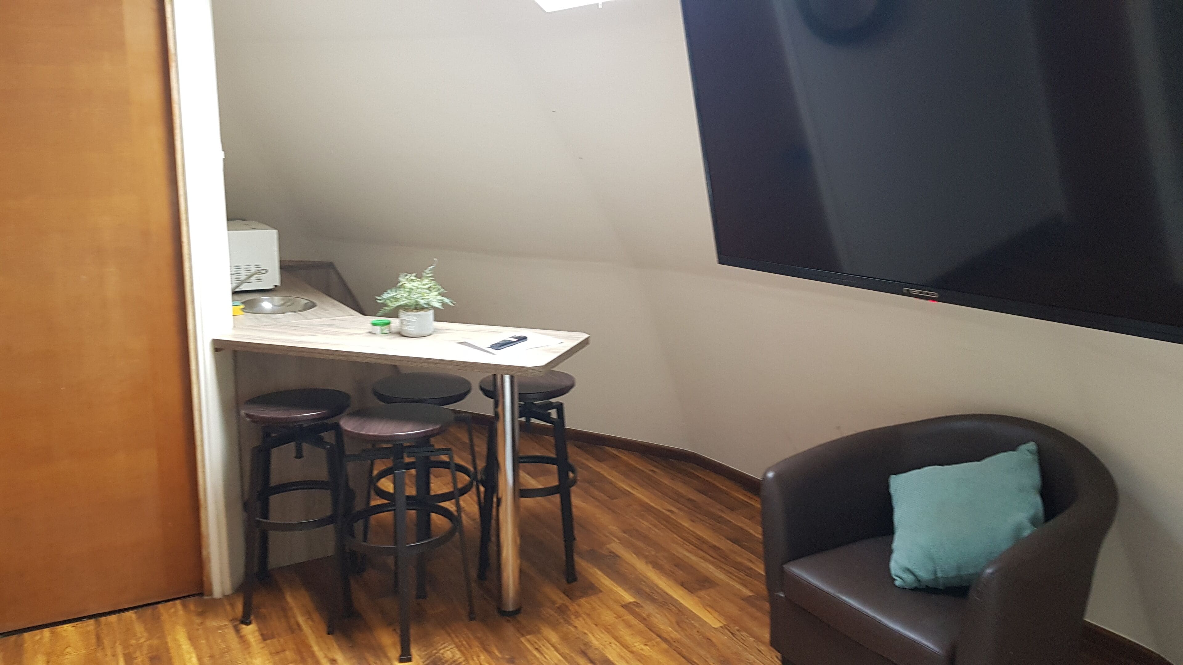 A compact room with a kitchenette, a dining area with stools, a single chair with a cushion, and a large TV mounted on the wall.