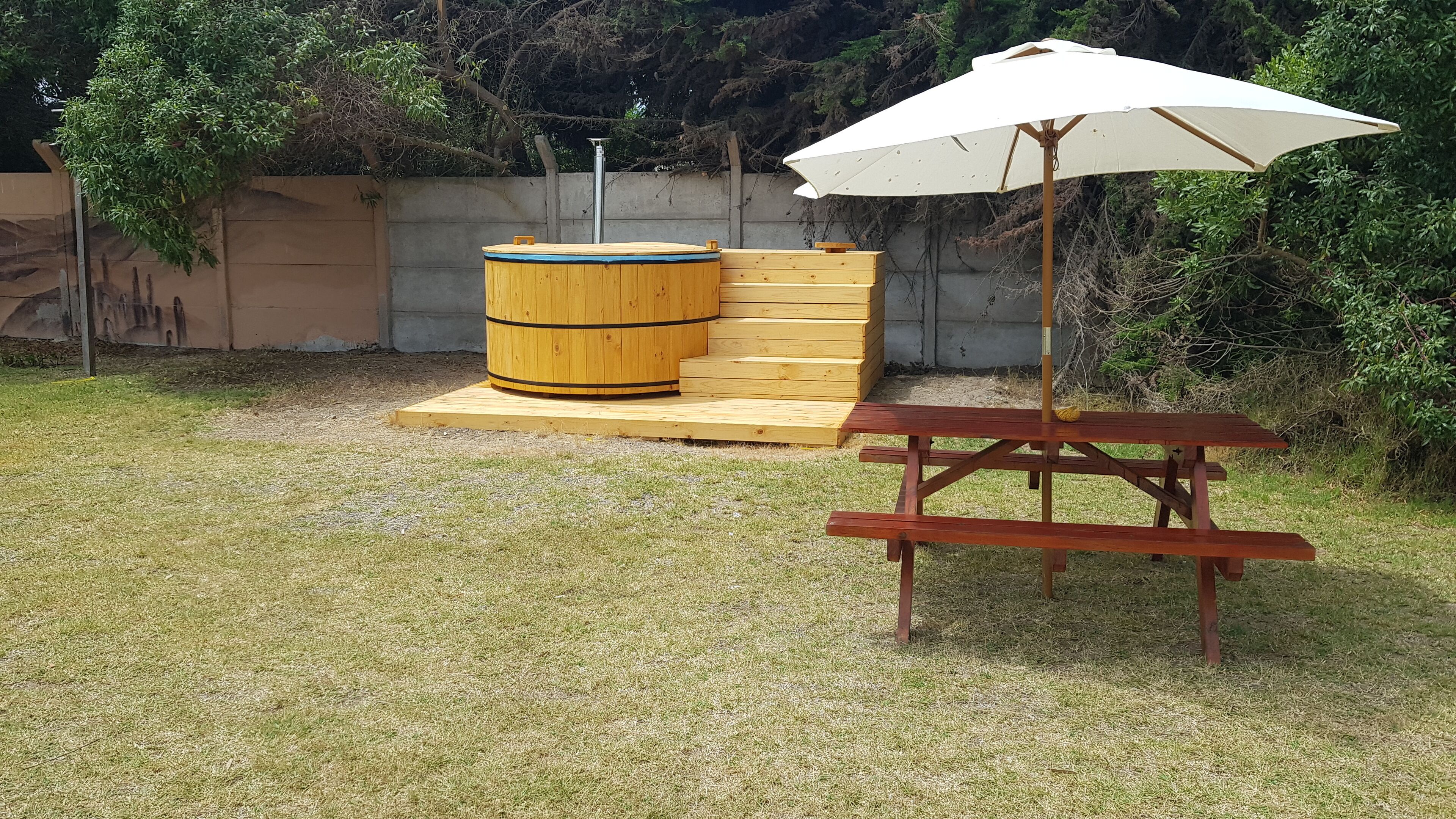 A wooden hot tub with steps, a picnic table, and an umbrella in a grassy backyard.