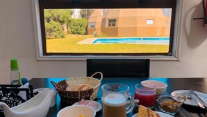 Una mesa de comedor con diferentes alimentos y bebidas, una ventana con vistas a una piscina y una casa, y una botella de agua.