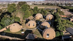 Un grupo de edificios con forma de cúpula rodeado de vegetación, con vistas a una ciudad y montañas lejanas.