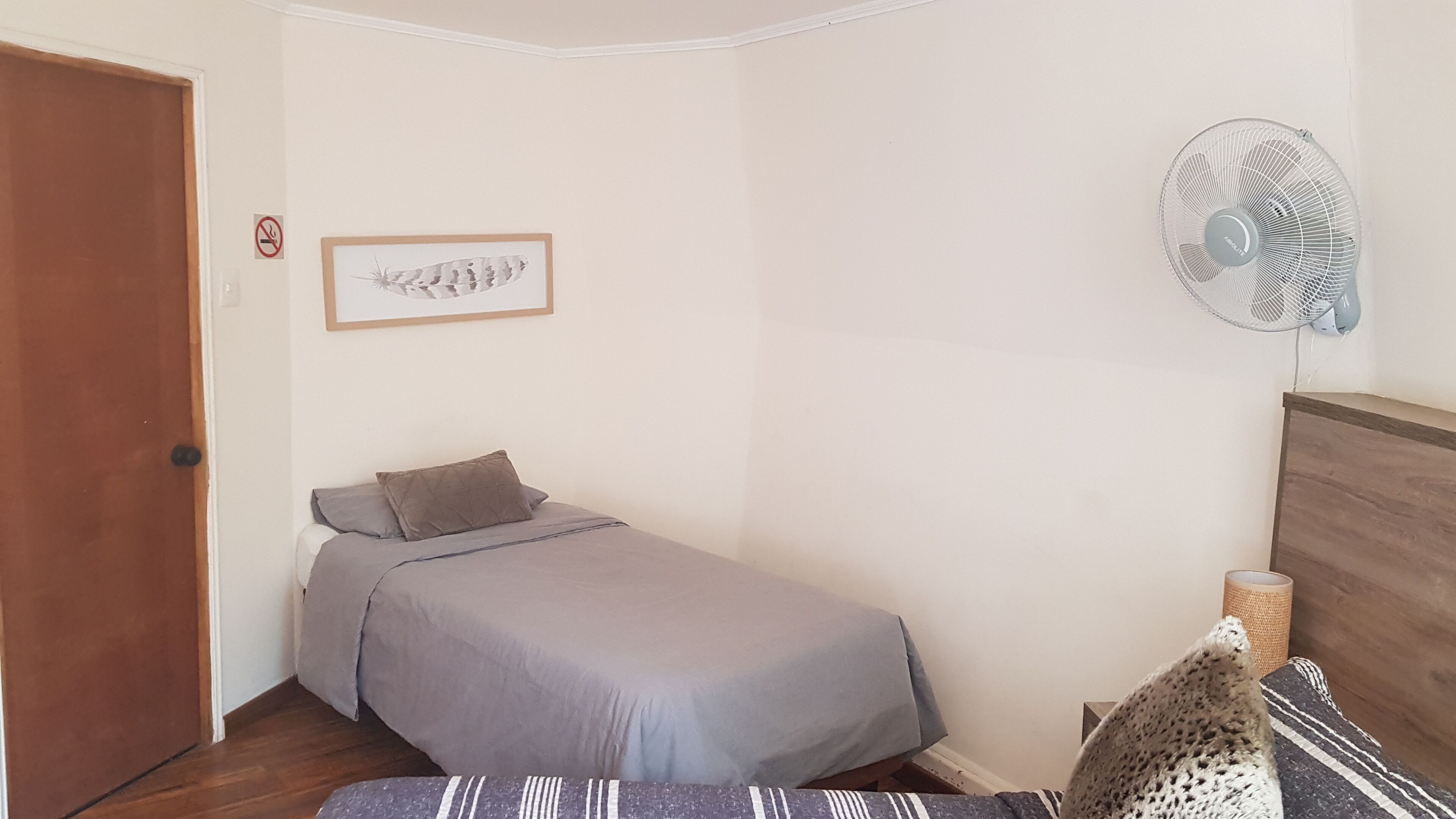 A small, neatly arranged bedroom with a single bed, a wall-mounted fan, and a framed picture.