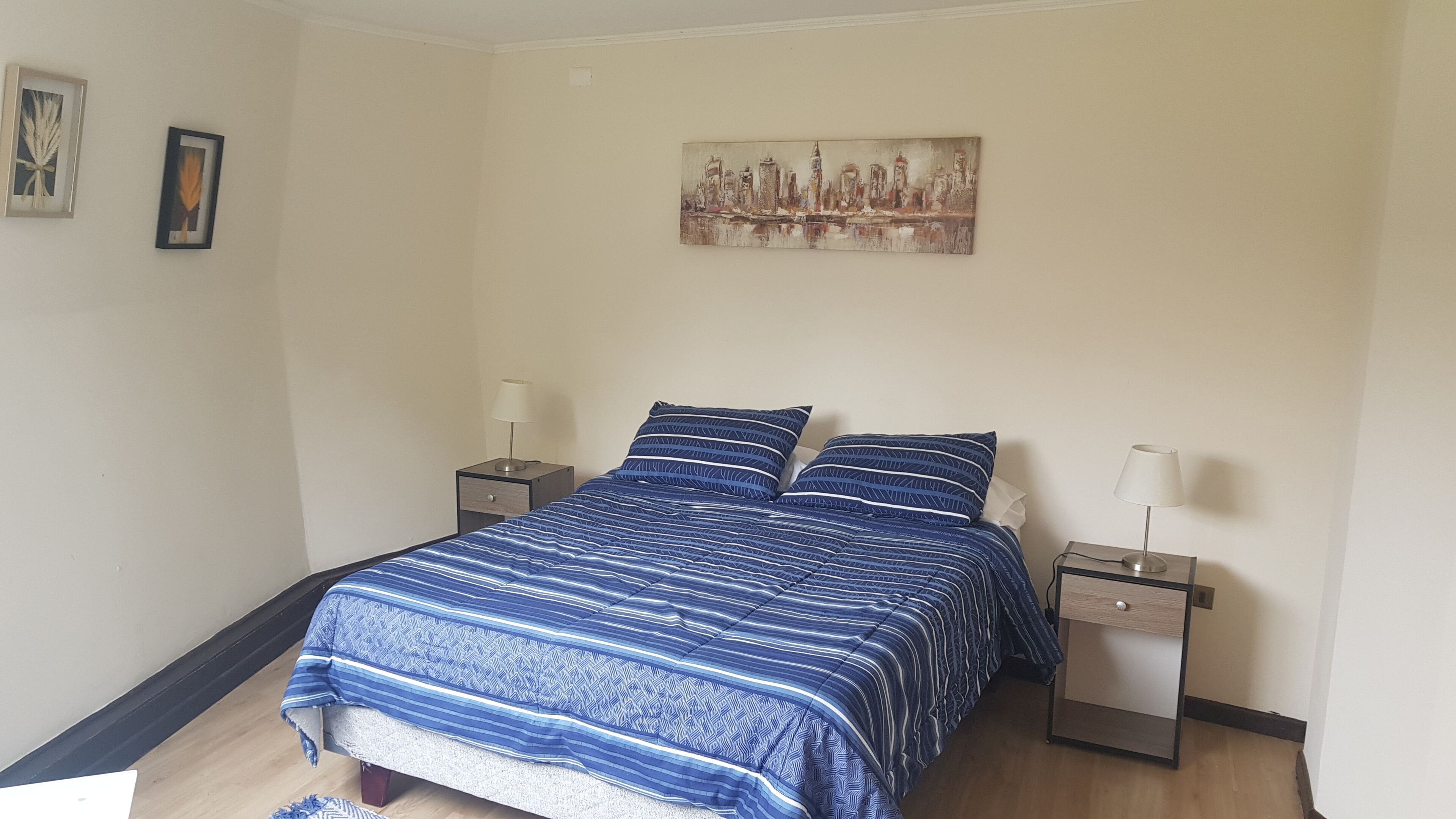 A bedroom with a bed, two nightstands, a lamp on each, a cityscape painting on the wall, and a small rug on the floor.
