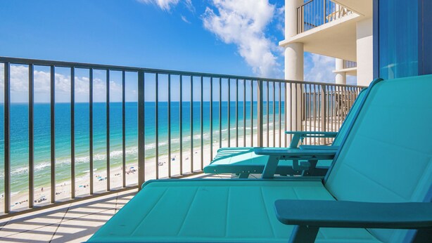 A balcony with a view of the beach and ocean, a pool area with lounge chairs, and a building with a balcony.