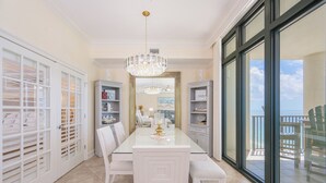 A spacious dining area with a glass table, white chairs, a chandelier, and a view of the outdoors.