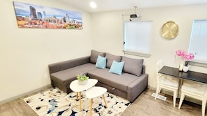 Ein Wohnzimmer mit einer Couch, einem kleinen Tisch mit Pflanze, einem Esstisch mit Stühlen, einer Uhr und einem Fenster mit Jalousien.