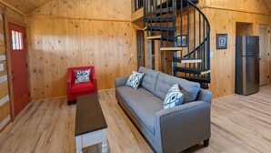 L’intérieur d’un chalet accueillant, avec un canapé gris, un fauteuil rouge, un plancher en bois, un escalier en colimaçon et un réfrigérateur.