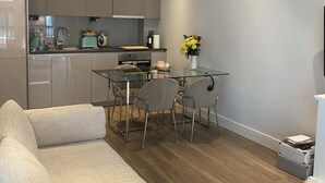 A modern living room with a grey sofa, a glass dining table with chairs, and a kitchen area with grey cabinets and a white countertop.