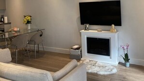 A modern living room with a flat-screen TV mounted above a fireplace, a glass dining table with chairs, and a cozy sofa.