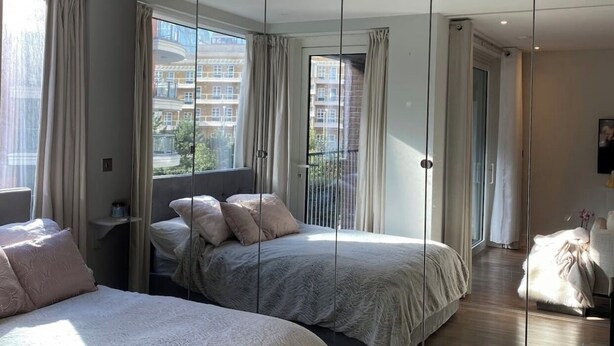 A modern bedroom with two beds, a large mirror, and a view of buildings through the windows.