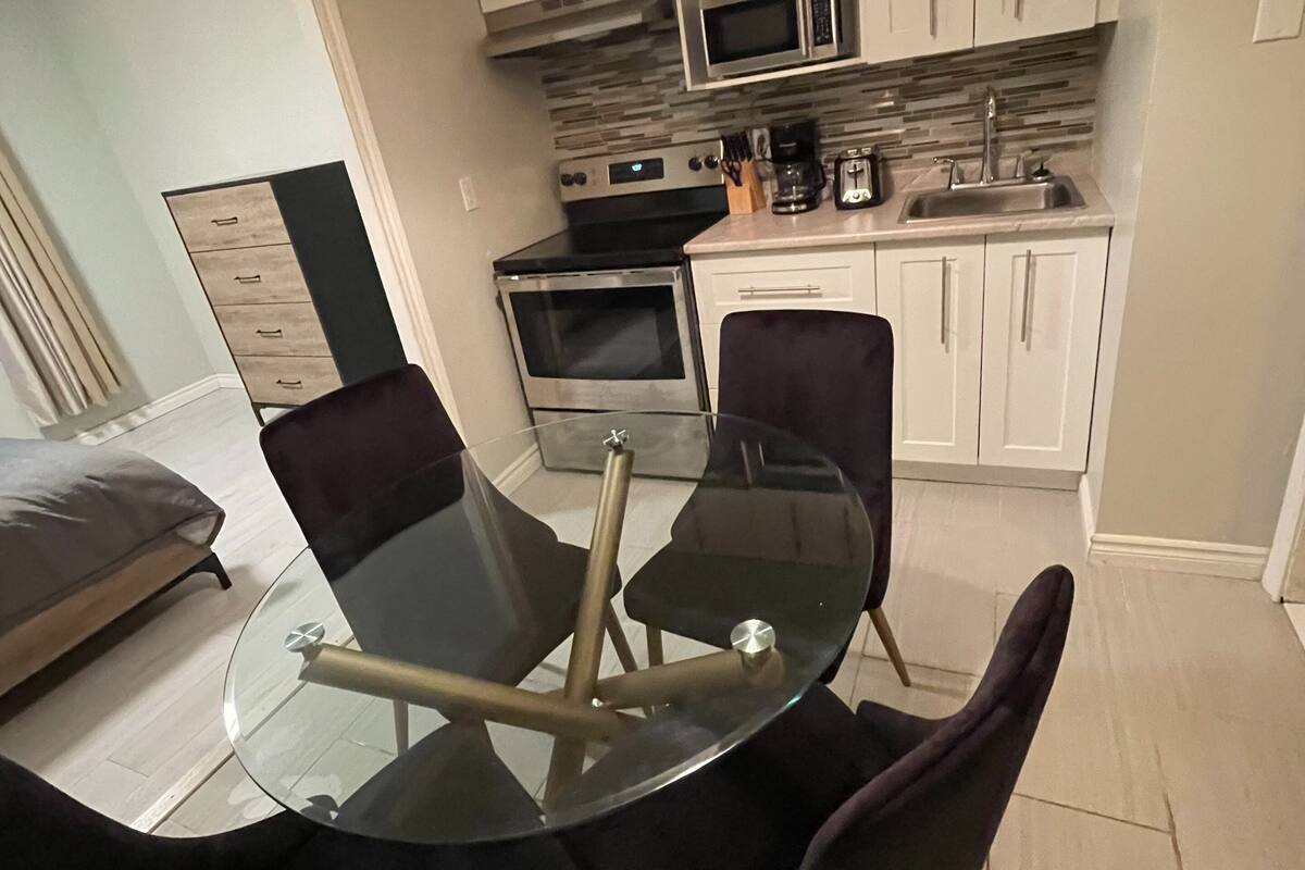 A modern kitchen with white cabinets, a microwave, oven, and a coffee maker. There is a glass dining table with chairs.