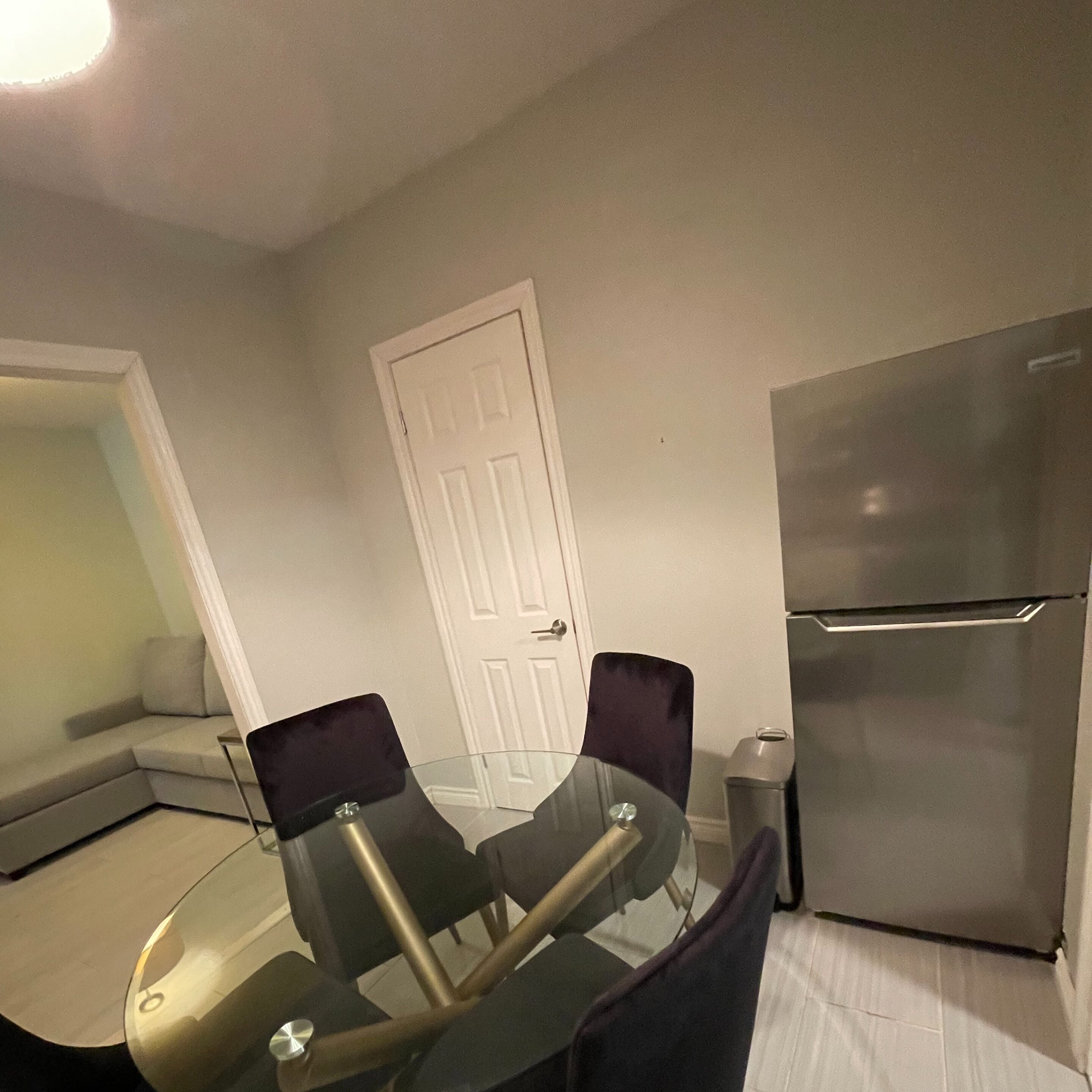 A compact kitchen with a glass-top dining table, two chairs, a white door, and a stainless steel refrigerator.