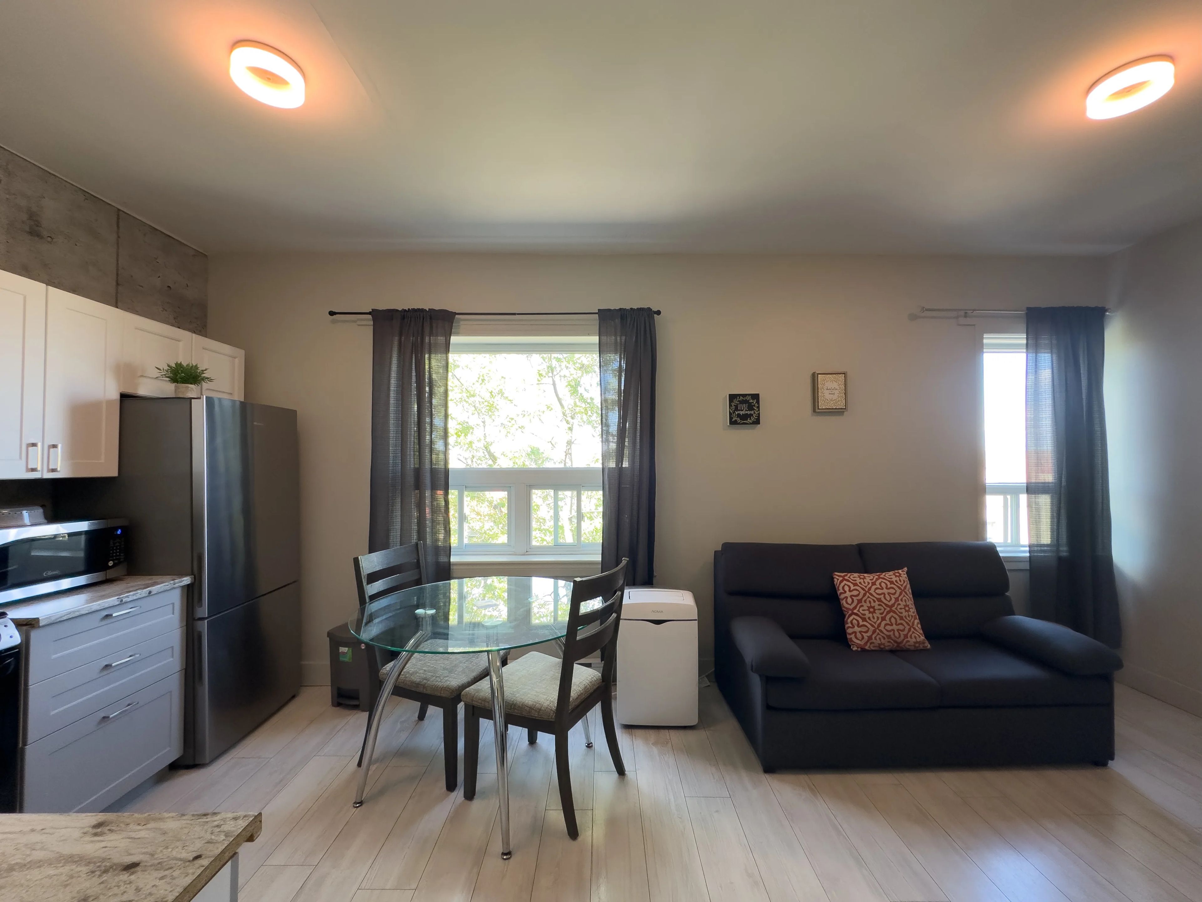 A modern kitchen with a refrigerator, microwave, and dining table with chairs. There is a sofa and a window with curtains.