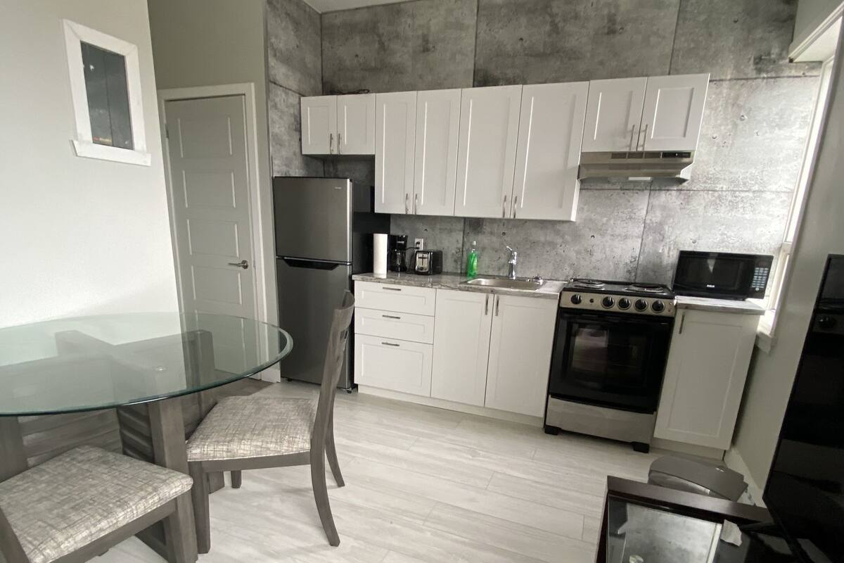 A modern kitchen with white cabinets, a stainless steel refrigerator, a gas stove, and a microwave. There is a glass dining table with two chairs.
