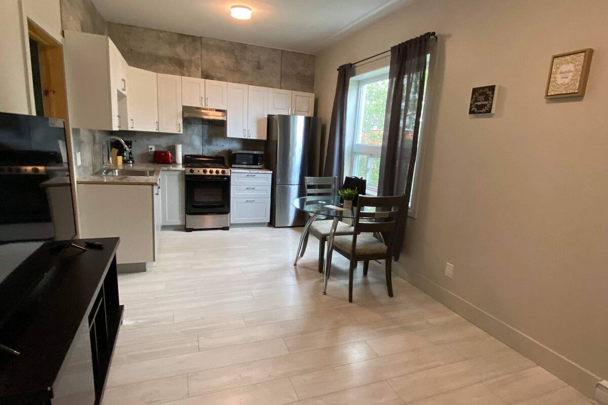 A modern kitchen with white cabinets, a stove, microwave, and refrigerator. There is a dining area with a table and chairs.