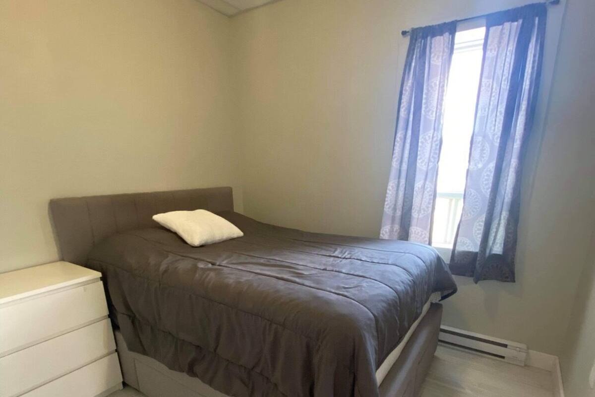 A small, neatly arranged bedroom with a bed, a bedside cabinet, and a window with curtains.