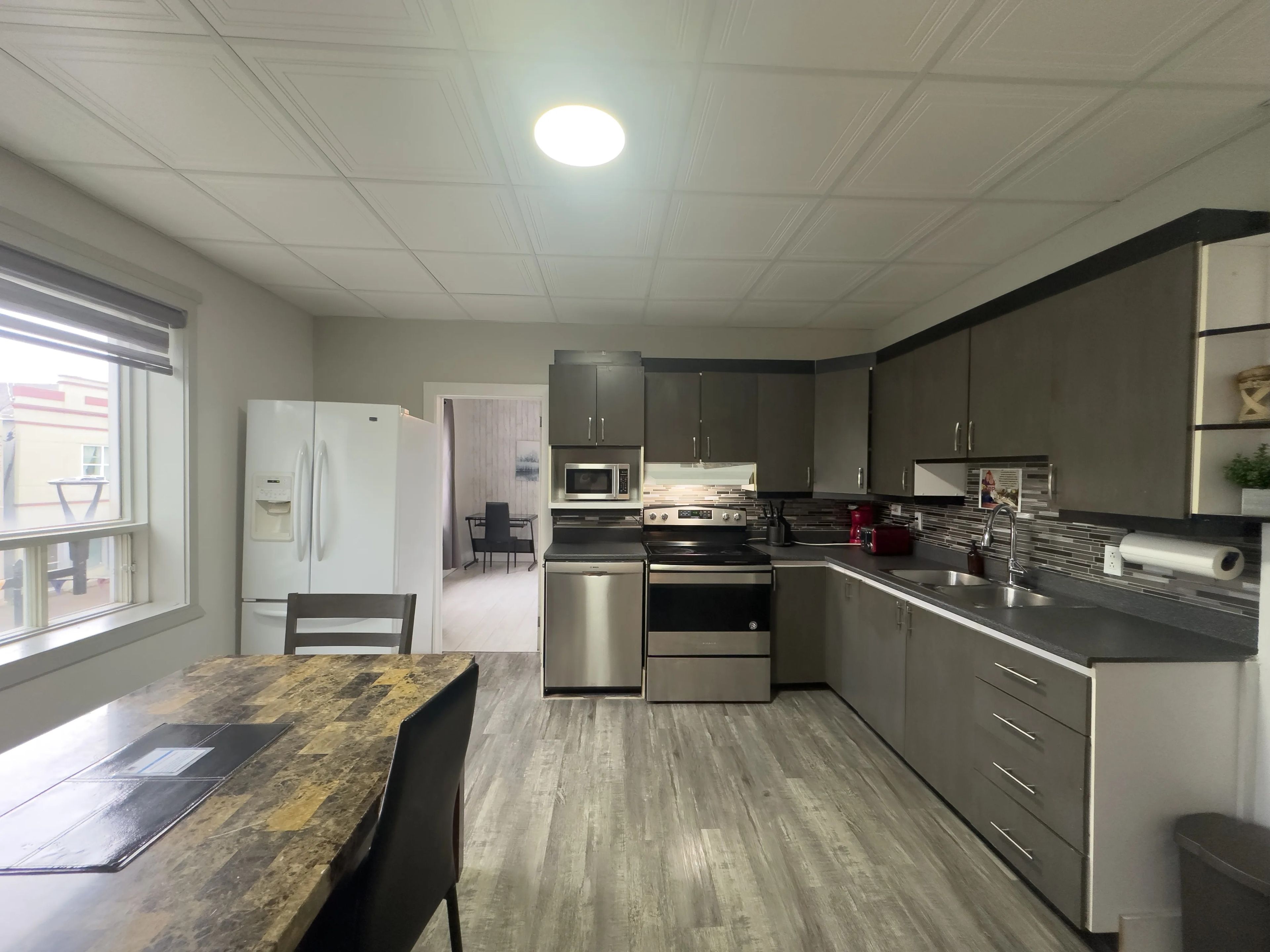 A modern kitchen with stainless steel appliances, a dining area with a wooden table, and a view through a window.