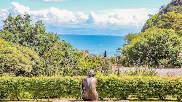 Une sculpture représentant une figure assise, surplombant un paysage côtier caractérisé par une végétation luxuriante et un ciel dégagé.
