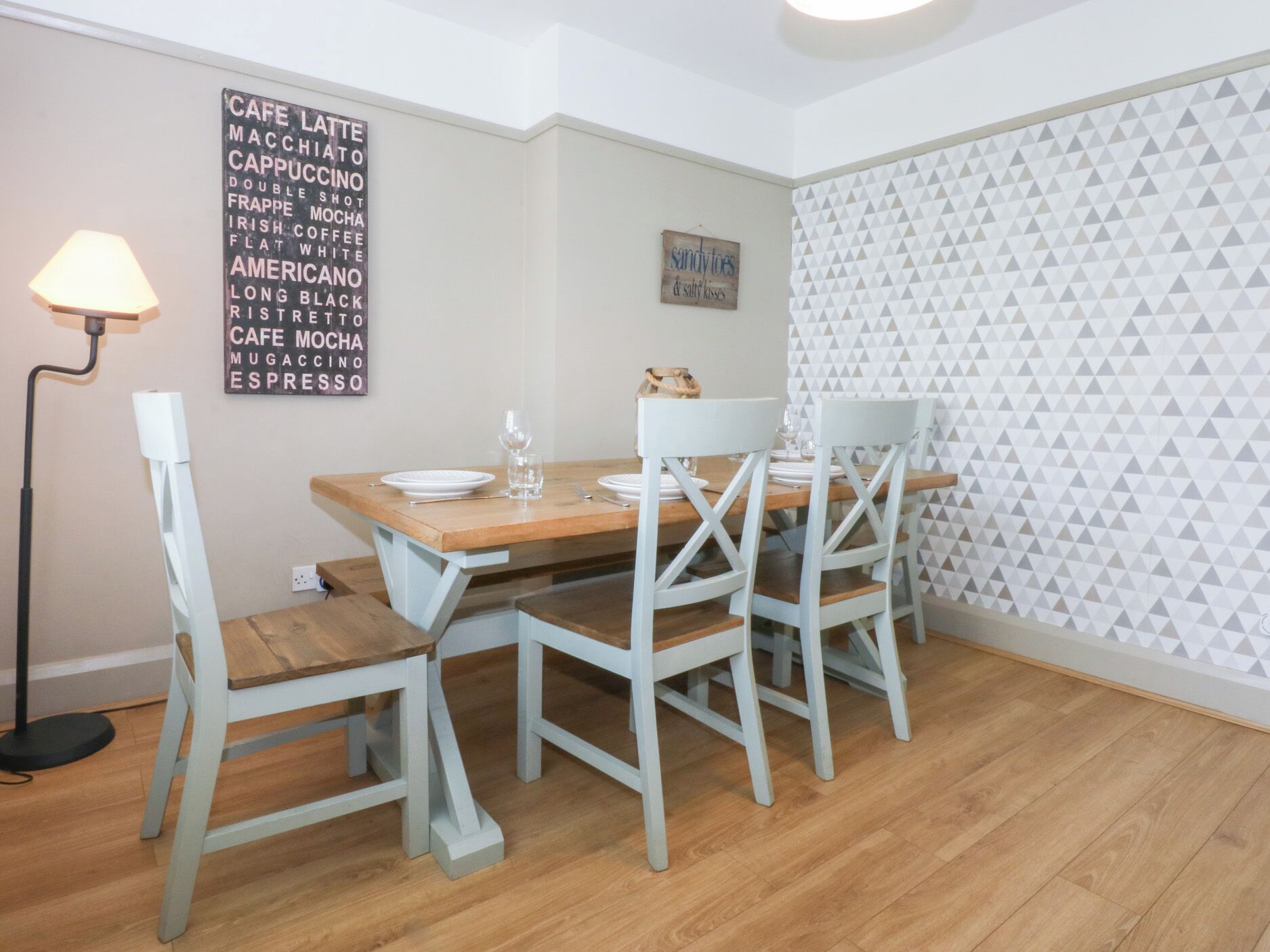 Un comedor con una mesa de madera y cuatro sillas, un letrero en la pared que enumera tipos de café, y una pared con un diseño geométrico.