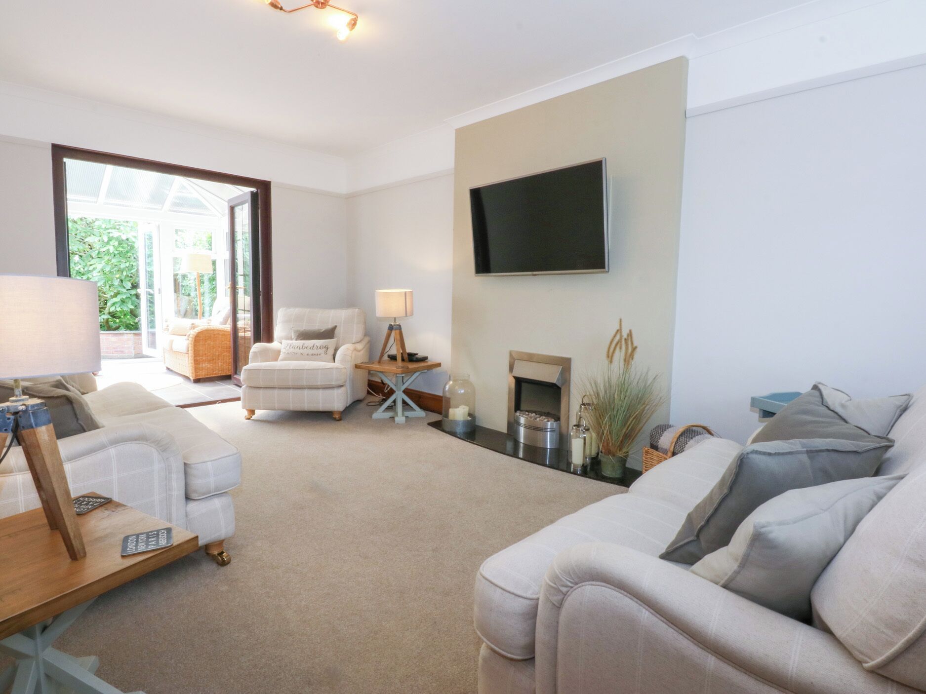 A living room with a sofa, armchair, and a small table with a lamp. There is a fireplace and a television mounted on the wall.