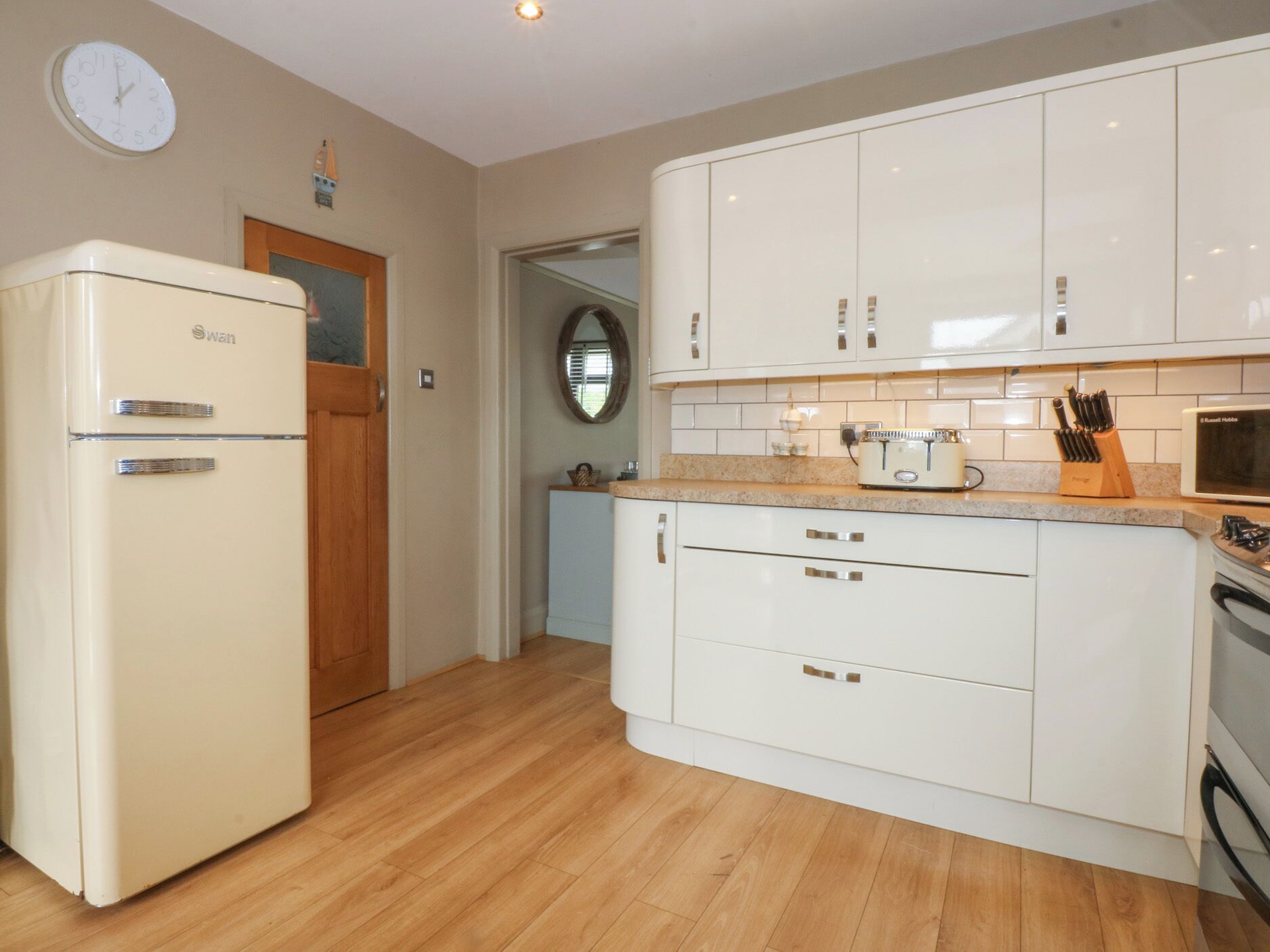Une cuisine avec des meubles blancs, un réfrigérateur Swanz d’époque, une porte en bois et un four à micro-ondes posé sur le plan de travail.