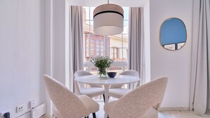 Un comedor moderno con una mesa redonda, dos sillas, una lámpara colgante y vistas a edificios a través de grandes ventanales.