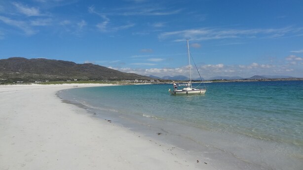 A beach with a sailboat anchored near the shore, clear turquoise water, and a distant mountain range.
