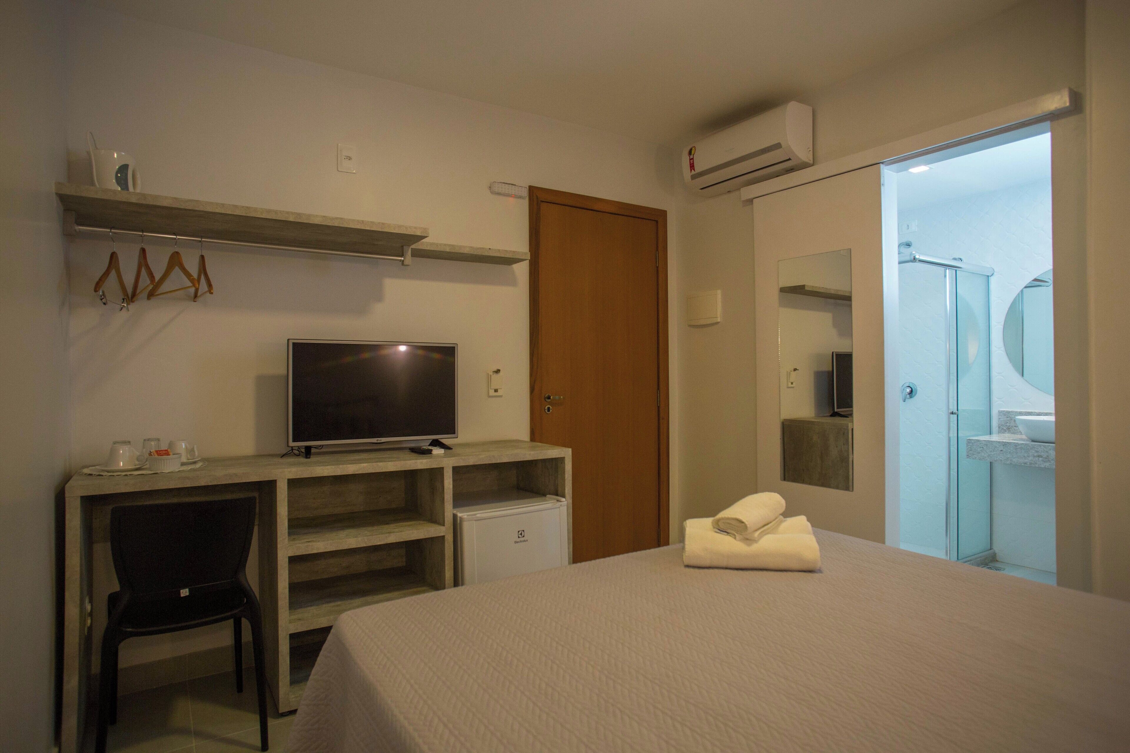 A hotel room with a bed, a desk with a television, a chair, a small refrigerator, a wooden door, and a bathroom with a glass shower.