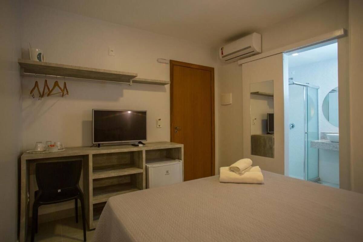 A hotel room with a bed, a desk with a television, a small refrigerator, a door, and a bathroom visible through an open door.