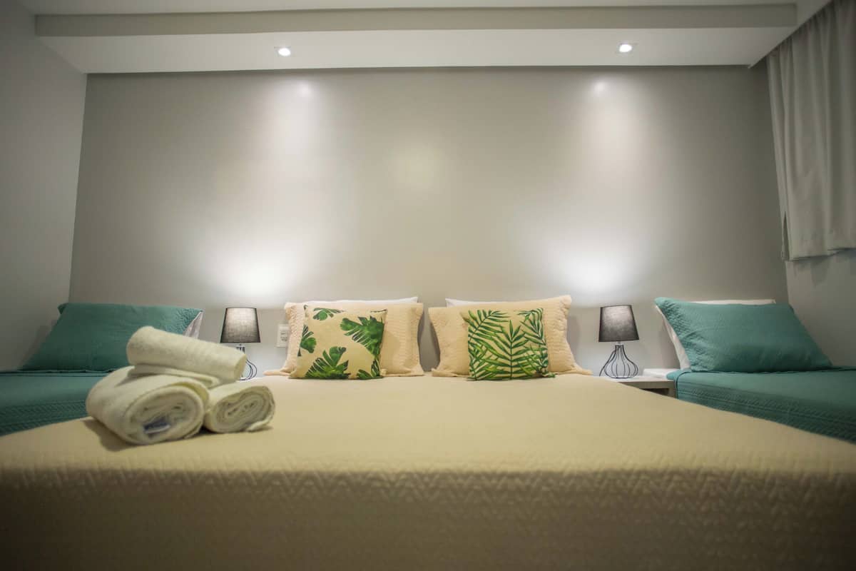 A double bed with a beige bedspread, two pillows with leaf patterns, a rolled towel, and two nightstands with lamps.