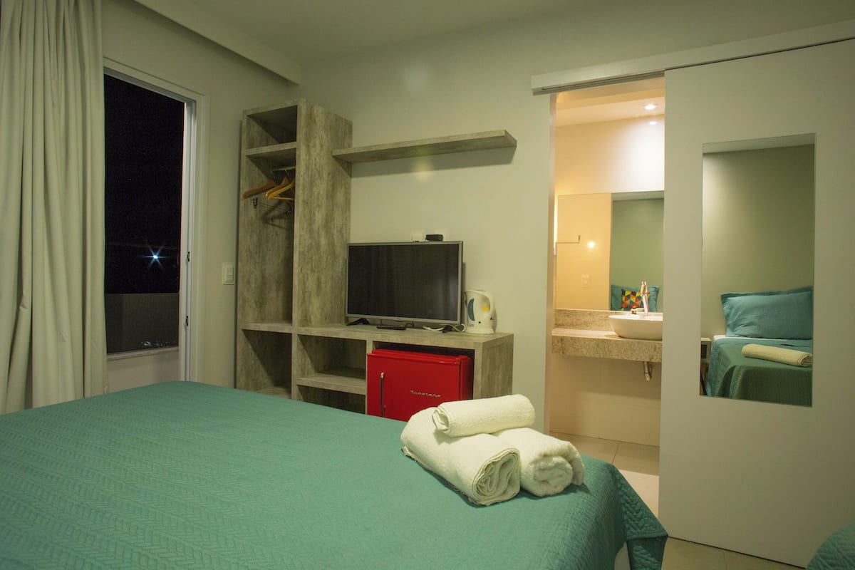 A hotel room with a bed, a TV, a red minibar, and a bathroom with a sink and mirror.