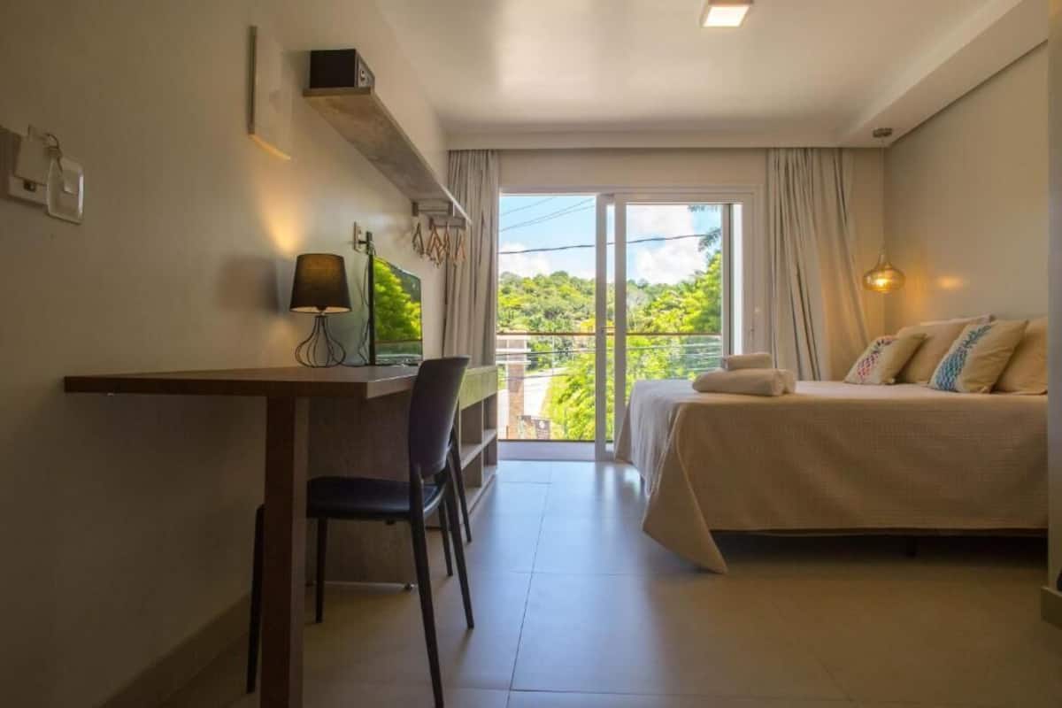 A hotel room with a bed, a desk with a chair, a TV, and a balcony with a view of greenery.