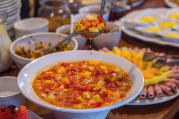 A bowl of vegetable soup with a spoonful being lifted, surrounded by various food items on a table.