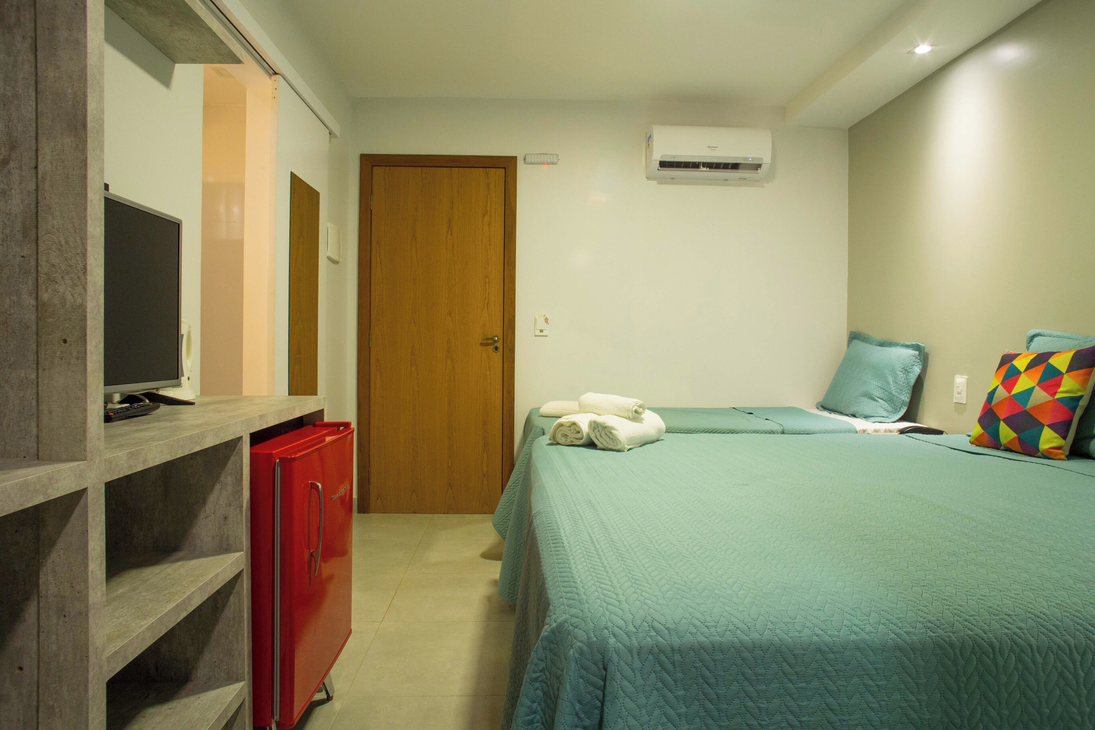 A bedroom with a bed, a red refrigerator, a wooden door, and an air conditioning unit.