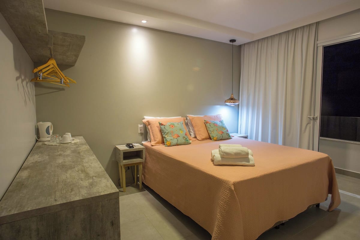 A modern bedroom with a large bed, a bedside table, a hanging clothes rack, and a window with curtains.