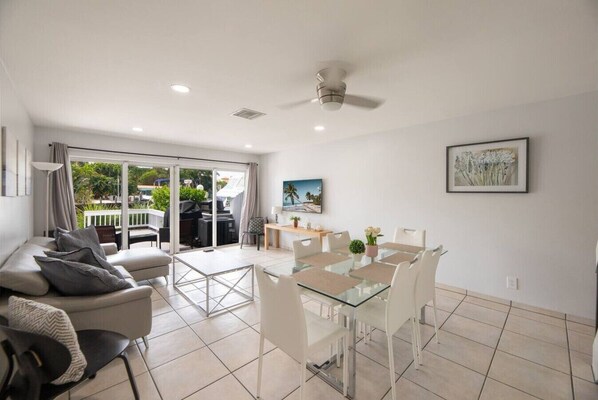 A modern living room with a glass dining table, white chairs, a sofa, and a ceiling fan.