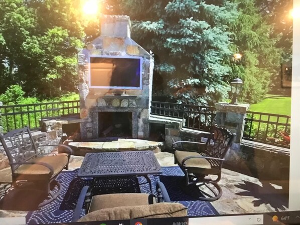 Relax by the stone-built fireplace, surrounded by lush greenery, with a flat-screen TV and comfortable seating for two.