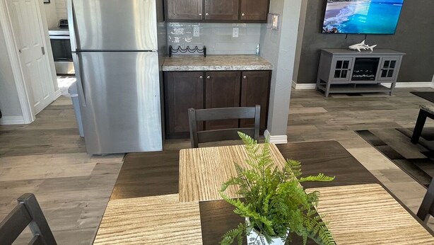 A modern kitchen with dark wooden cabinets, a stainless steel refrigerator, and a countertop with decorative items. There is a dining table with a plant centerpiece and a TV mounted on the wall. - Elephant Lake Resort Oasis New (Elephant Butte)