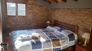 A bedroom with a brick wall, a wooden bed frame, a quilted bedspread, and a window with frosted glass. - Cabin Arroyo de Uco - Between mountains, streams and wineries (Manzano Historico)