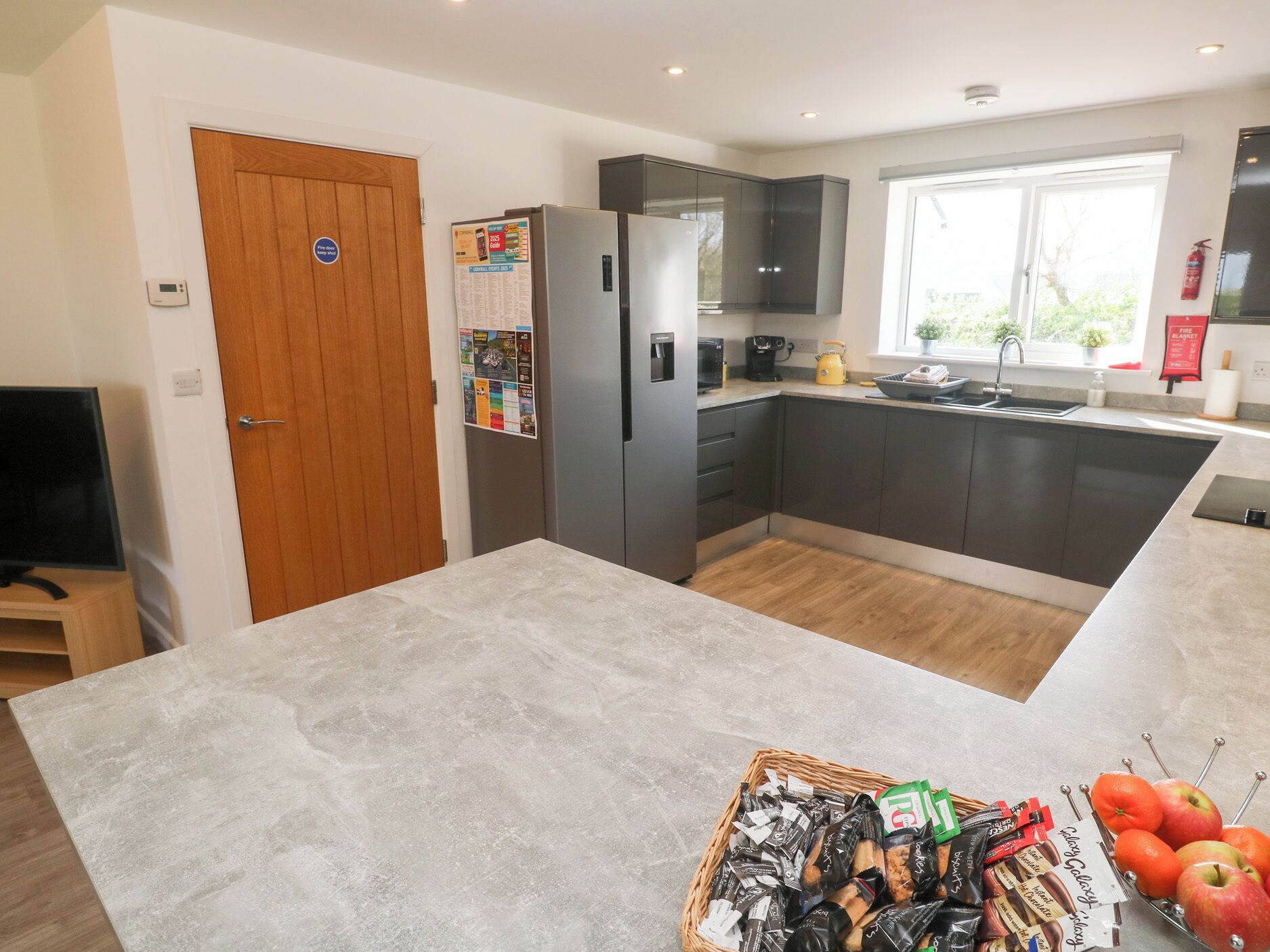 A modern kitchen with grey cabinets, a built-in oven, and a large island with a basket of snacks and fruits.