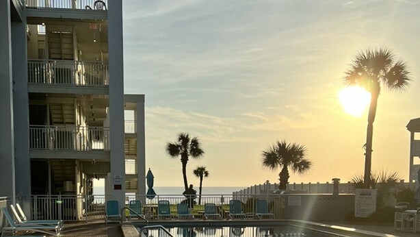 Un espace aménagé au bord de la piscine avec des chaises longues, un bâtiment doté de balcons et des palmiers.
