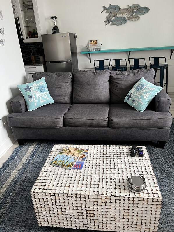 A grey sofa with two decorative pillows, a woven coffee table with magazines and remotes, and a kitchen area in the background.
