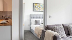 Ein modernes Schlafzimmer mit grauem, gepolstertem Kopfteil, einer weißen Bettdecke und einer grauen Couch mit flauschigen Kissen.