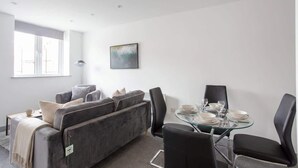 Ein modernes Wohnzimmer mit einem grauen Sofa, einem Glastisch für vier Personen und einem großen Fenster, das natürliches Licht hereinlässt.